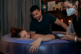 Photo captures dad supporting mom as she labors during childbirth at a home birth; Man applies counterpressure to womans back during a contraction