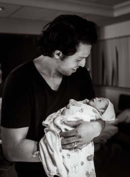 Photo of dad smiling while holding newborn baby in hospital birth setting