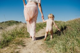 Image of toddler picking at grass while holding mother's hand. Mother is pulling up a long pink skirt as she walks