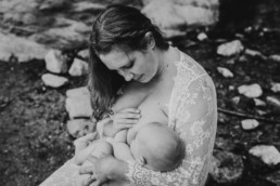 Black and white image of mother looking down at her baby as she breastfeeds. They sit on a rock next to a river