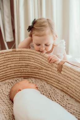 Newborn baby lays in moses basket while toddler sister peers down at him from outside the basket