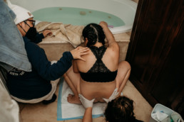 Laboring women is in a squat leaning against a tub. A midwife applies pressure on her hips while another midwife applies gentle touch to woman's shoulder.