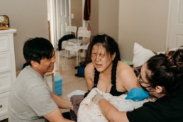 Women has a look of exasperation and relief immediately following the birth of her baby at a homebirth. Father is looking at her smiling. Midwife is holding a towel against the baby.