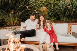 Family of three sit on a white couch outside while a dog is at their feet. They are in front of a fire pit. Mom and daughter drink from mugs. Dad and mom look at daughter.
