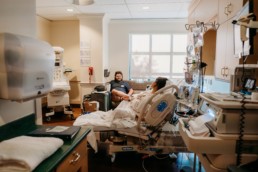 Pull back image of laboring mother in hospital bed looking at father who is sat on the couch smiling at her. Image contains hospital medical equipment meant for birth.