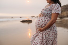 Pregnant woman holds her belly with sun setting behind her. She stands on a shoreline gazing out to the water.