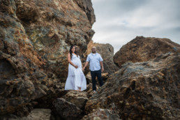 Pregnant woman stands next to partner in dramatic rock scene while holding hands. Both look at camera