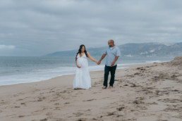 Pregnant woman walks with partner along beach. Women holds her belly and looks downward