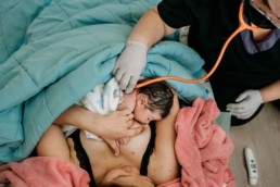 Mother hold newborn baby during homebirth while midwife listens to baby with stethoscope