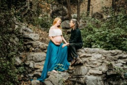 Woman and man look at each other lovingly. Mother is holding her pregnant belly. Couple sits on a wall in the woods