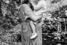 Black and white image of mother holding toddler over her pregnant belly. The pair stand in front of a waterfall in the woods.