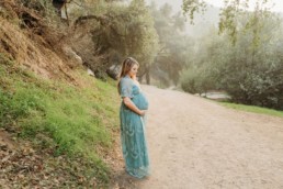 Pregnant woman stands next to green hill in the forest while gazing downward and holing her belly.