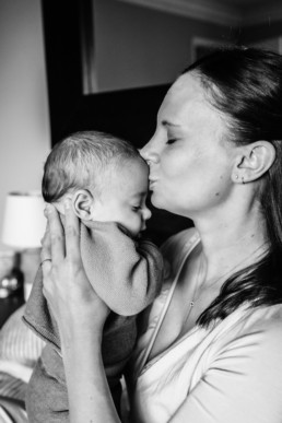 Black and white image of mom holding baby up to her lips to kiss baby on the forehead. Both have their eyes closed.