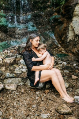 Mother sits at foot of a waterfall and breastfeeds toddler while holding him close. She has her eyes closed and looks peaceful