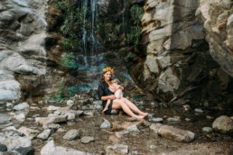 Mother sits at foot of a waterfall and breastfeeds toddler while holding him close. Mother is wearing a flower crown and smiling at the camera