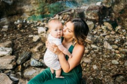 Mother sits holding baby amongst a riverbed. Mother kisses baby on the cheek while baby looks at the camera