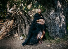 Pregnant woman leans against a tree in a forest while dressed in black tulle. She gazes upwards while resting hands on her belly