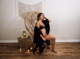 Pregnant woman perches on edge of peacock chair, She is surrounded by candles and draped in black fabric